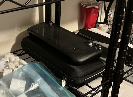 A Steam Deck sitting on top of its carrying case on a shelf of miscellaneous tech junk.
