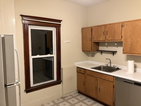 A bare kitchen with laminate counters, cheap cabinets, and bad stick-on tile flooring. Nice woodwork on the window though.