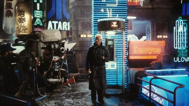 color production still from Blade Runner (1982) showing Rutger Hauer as Roy Batty in a black trenchcoat on a rain-soaked street full of neon signs. Film cameras and crew off to the left. Among other corporate signage in the background, a big Atari logo.