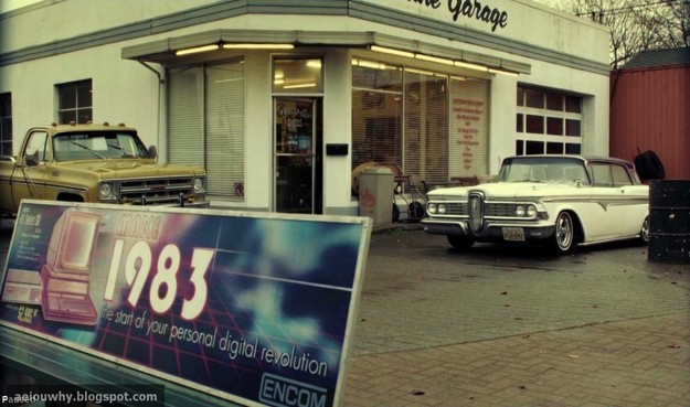 A scene from the TV show "Once Upon a Time", showing a gas station with a couple of 1980s cars. In the foreground is a billboard advertising an Encom computer: "Make 1983 the start of your personal digital revolution."