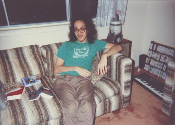 Zarf, age about 22, sitting on a rather nice striped couch. I'm wearing a vivid green t-shirt (it says "Musikanten" although you can't read that). A couple of books are lying next to me, along with a very 90s boombox (just visible). In the corner of the room is a rack of cassette tapes and a (musical) electric keyboard. There's also an abstract ceramic art thing next to the couch.