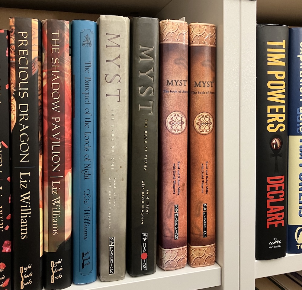 A closeup of a bookshelf. All three Myst novels are visible in the Hyperion hardcover editions. To the left is Liz Williams; to the right is Tim Powers. No, I don't shelve in alphabetical order.